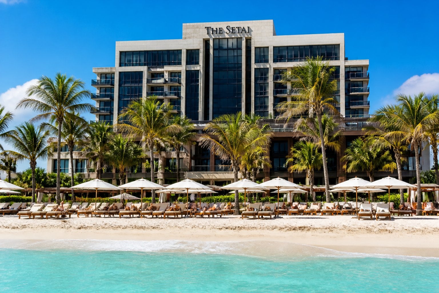 The Setai hotel in Miami Beach with palm trees, white sandy beach, turquoise ocean, and sun loungers under a clear sky.