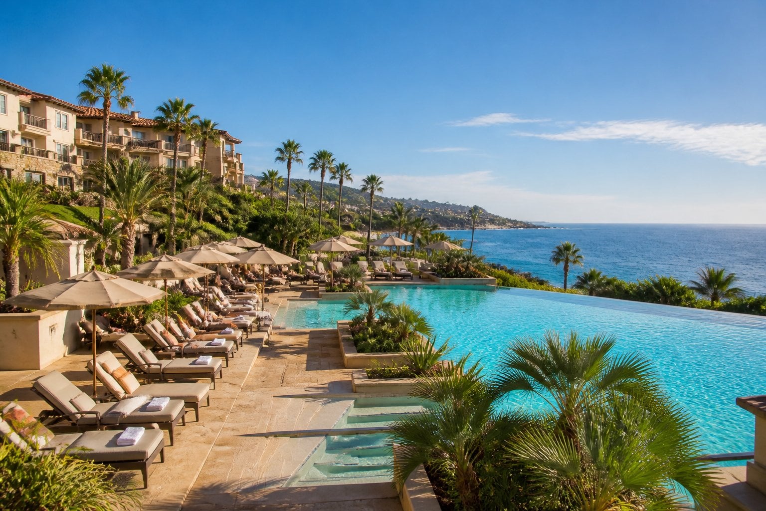 Oceanfront luxury resort with a swimming pool, palm trees, and Mediterranean-style buildings overlooking the Pacific Ocean.