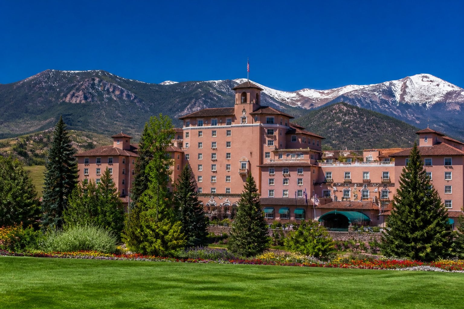 The Broadmoor hotel with stone architecture, surrounded by green gardens and pine trees, set against snowy mountain peaks under a blue sky.
