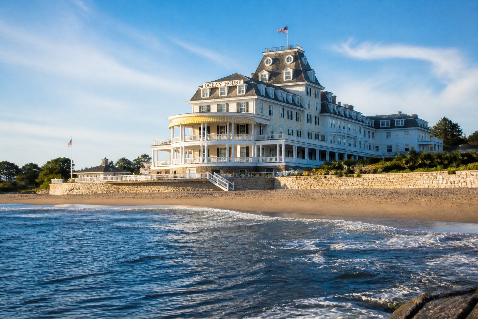 Ocean House hotel by the beach with white Victorian-style architecture, sandy shore, and calm ocean waves under a clear sky.