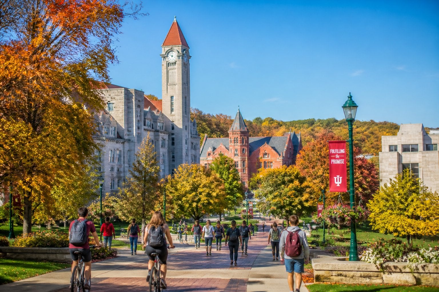 Students walking and biking on tree-lined paths near red brick buildings on a sunny day in Bloomington, Indiana, with autumn trees and rolling hills in the background.