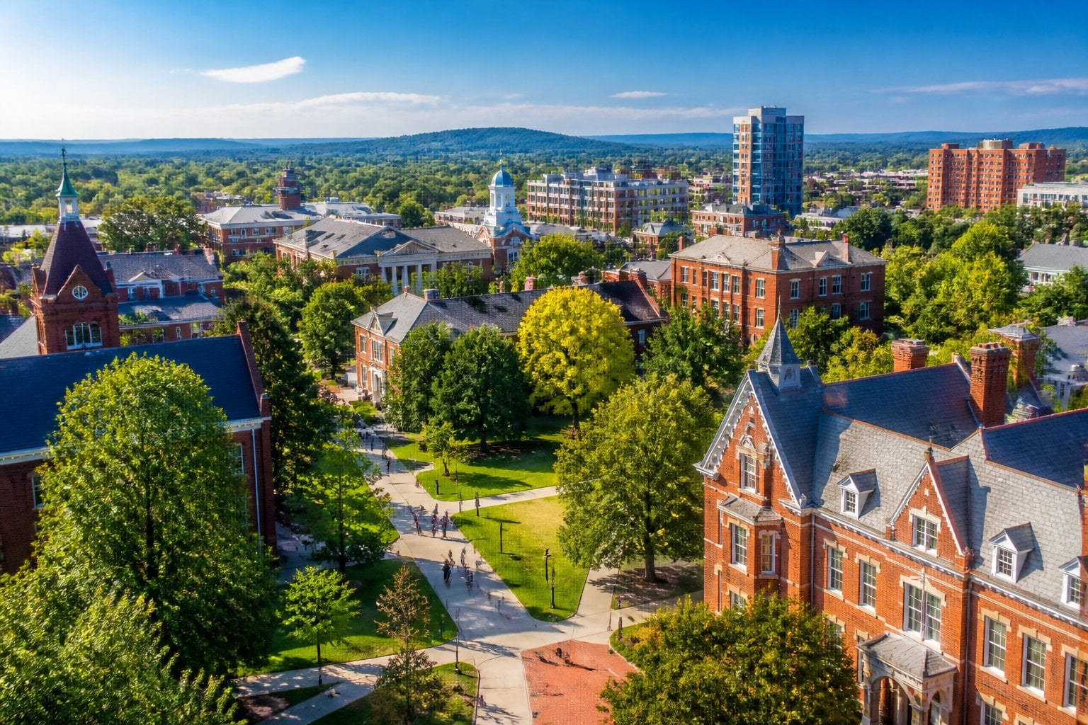Aerial view of Lexington, Kentucky college town with university buildings, green trees, students walking and biking, and rolling hills in the background.