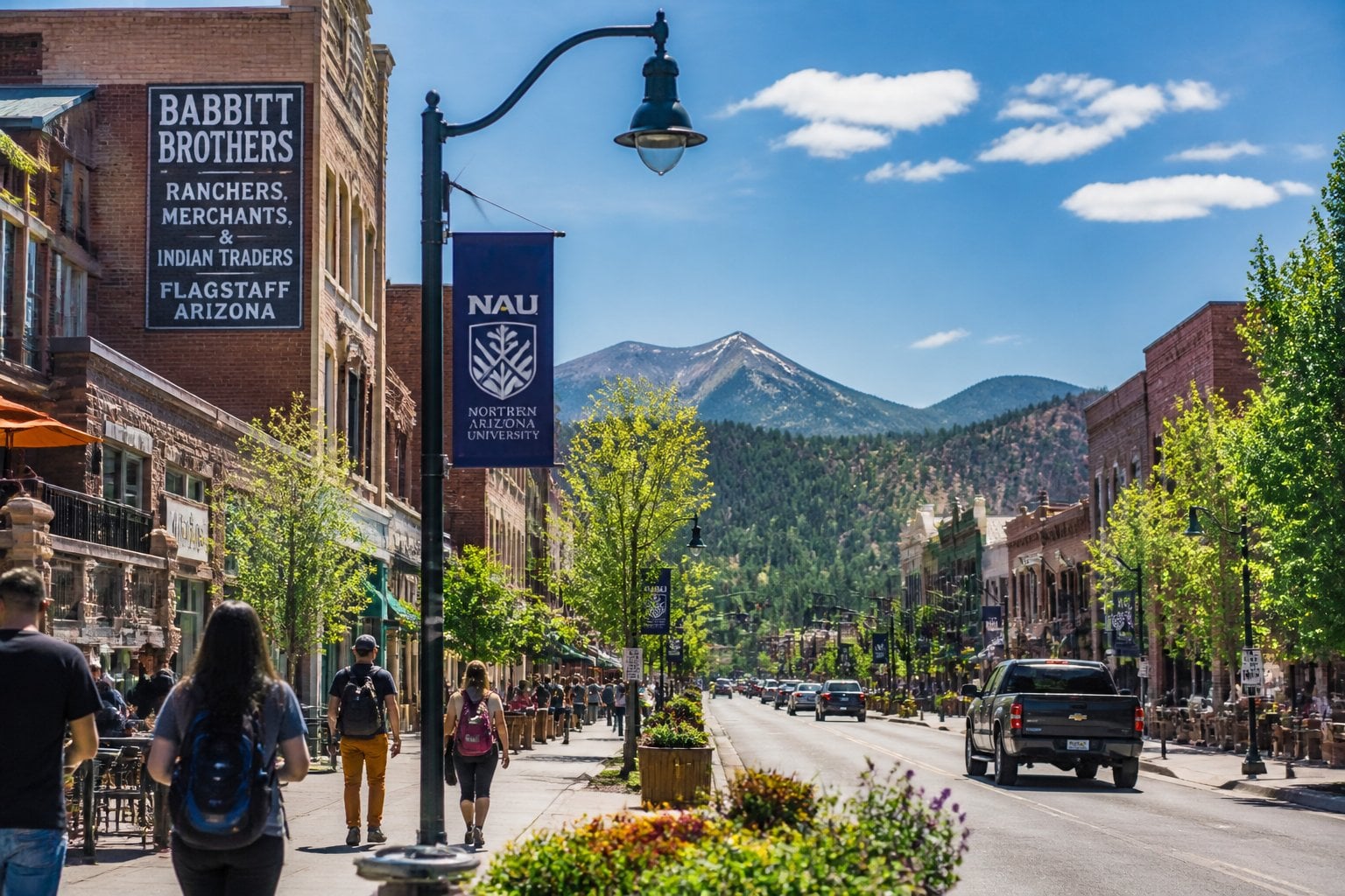 A busy downtown street in Flagstaff, Arizona, with people walking, outdoor cafes, pine trees, and mountains in the background.