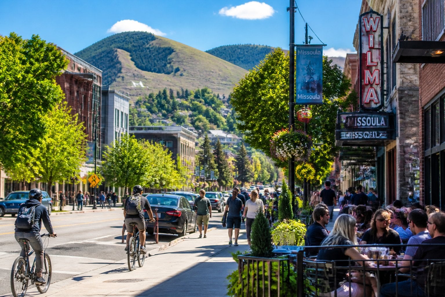 Downtown Missoula, Montana with people walking near shops and cafes, surrounded by trees and mountains in the background.