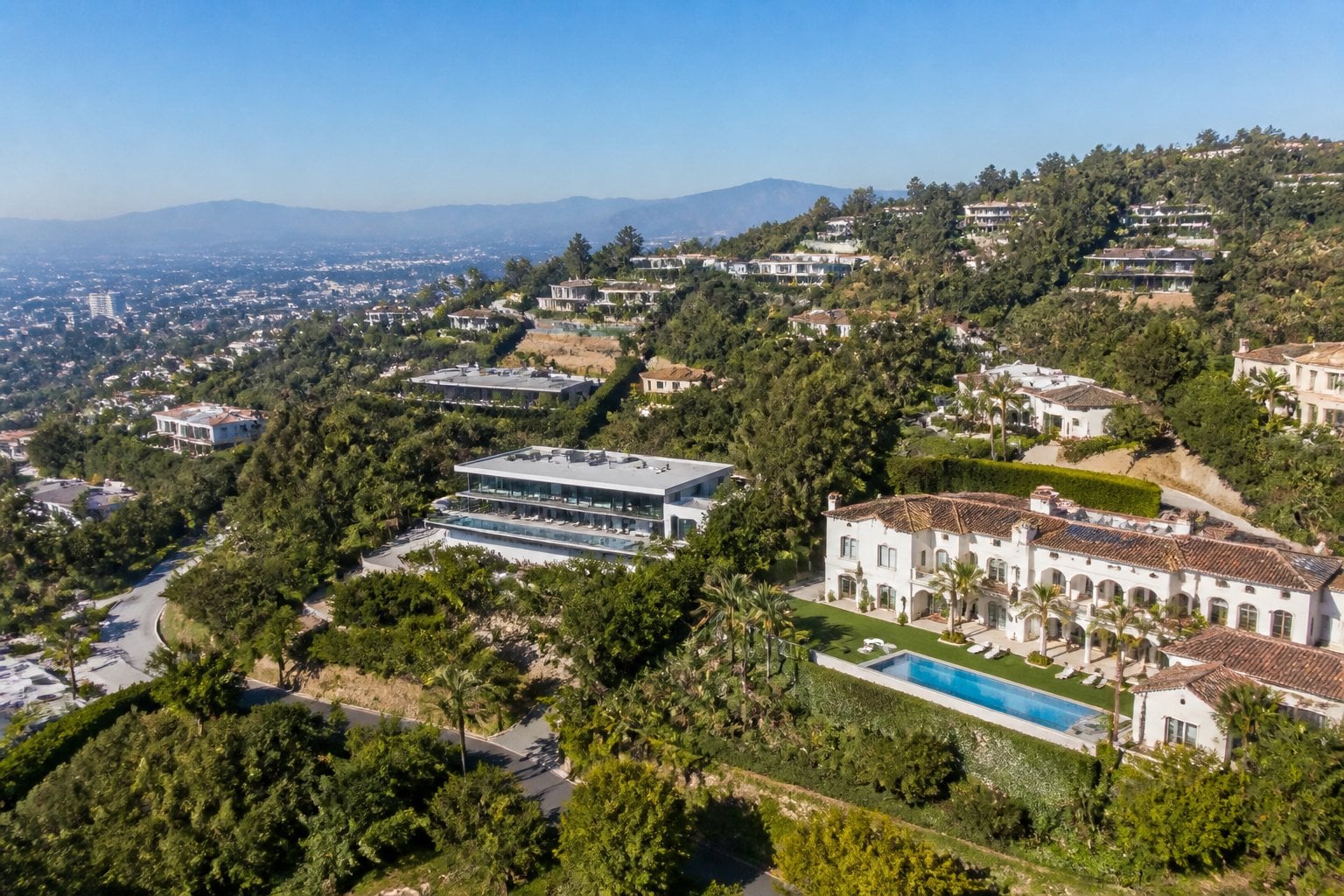 Aerial view of large luxury homes surrounded by trees and hills in a wealthy residential neighborhood.