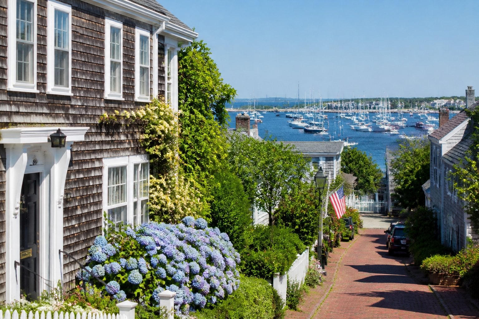 A coastal town with historic shingled houses, colorful gardens, and sailboats in a calm harbor under a clear sky.