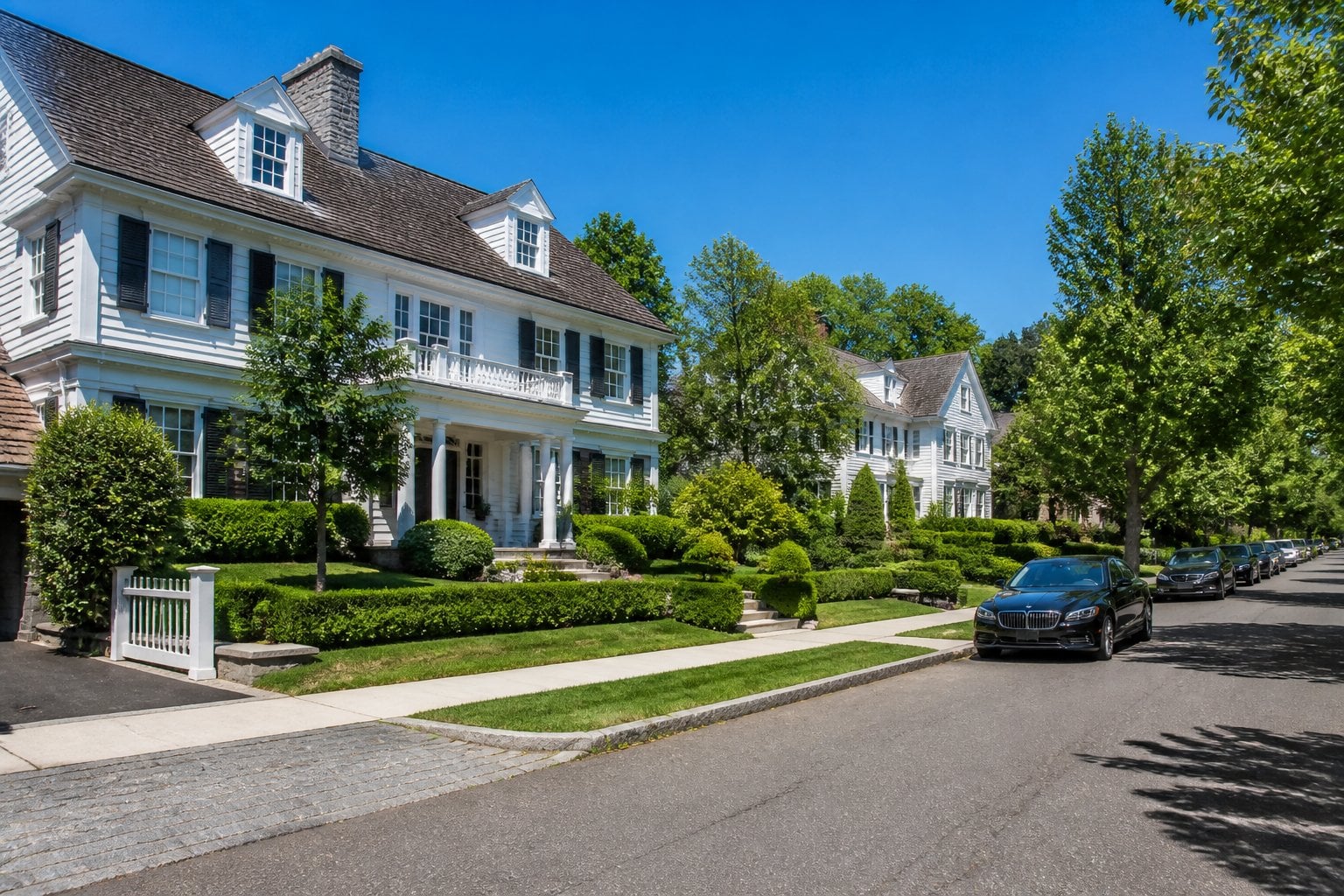 A quiet street in a wealthy neighborhood with large houses, green lawns, and trees under a clear blue sky.