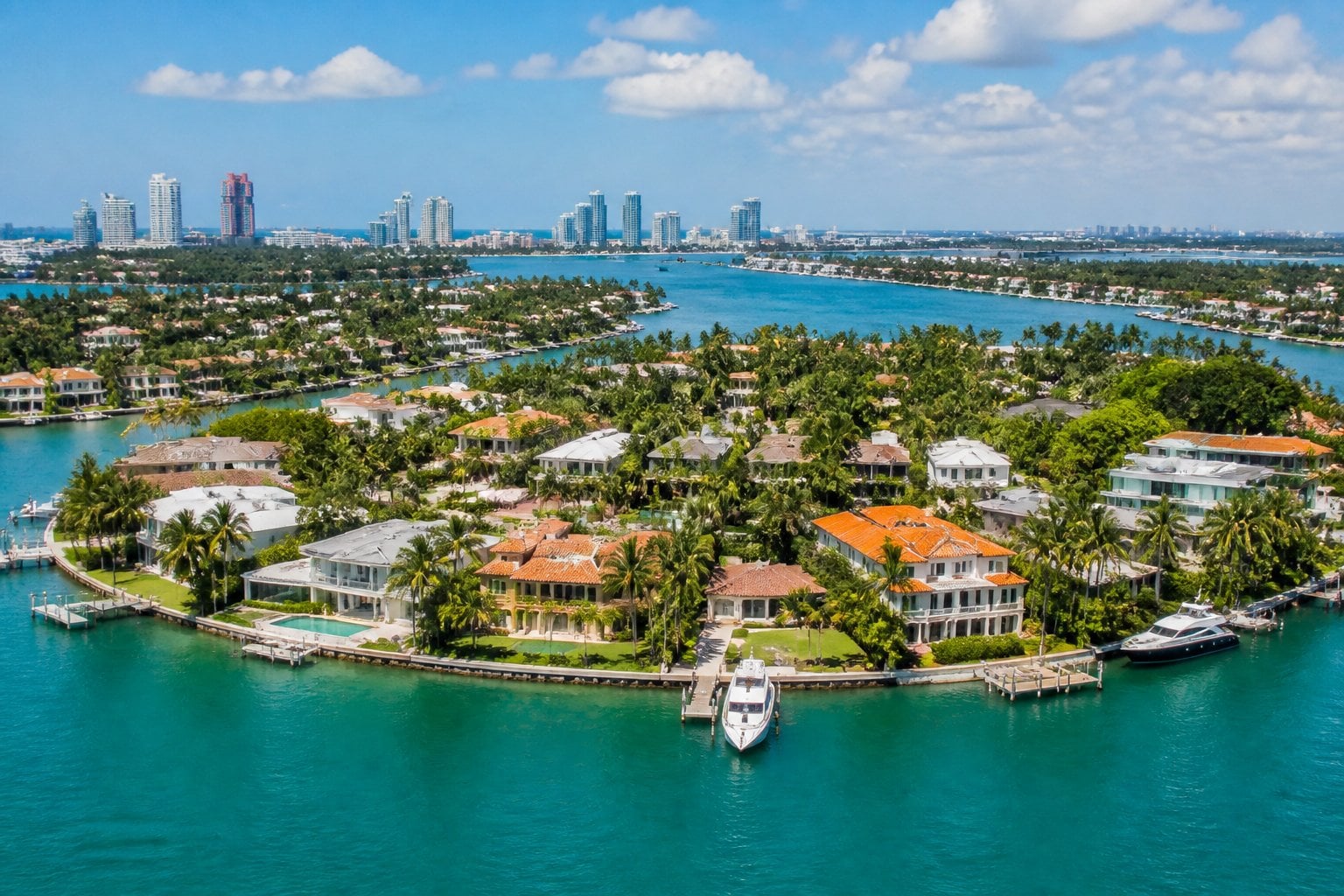 Aerial view of luxurious waterfront mansions with private docks and yachts on Star Island, Miami Beach, surrounded by clear blue water and palm trees.