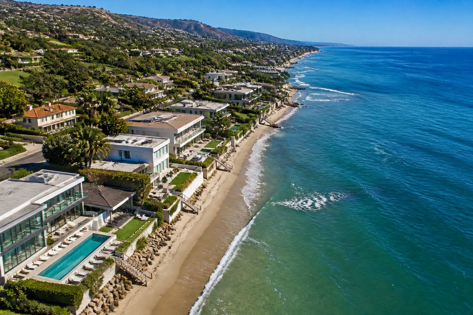 Aerial view of luxury beachfront homes along the coast of Malibu with the ocean and sandy beach in the background.