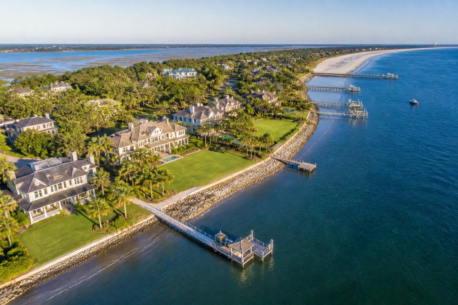 Aerial view of a coastal neighborhood with large waterfront mansions, green lawns, palm trees, and blue water under a clear sky.