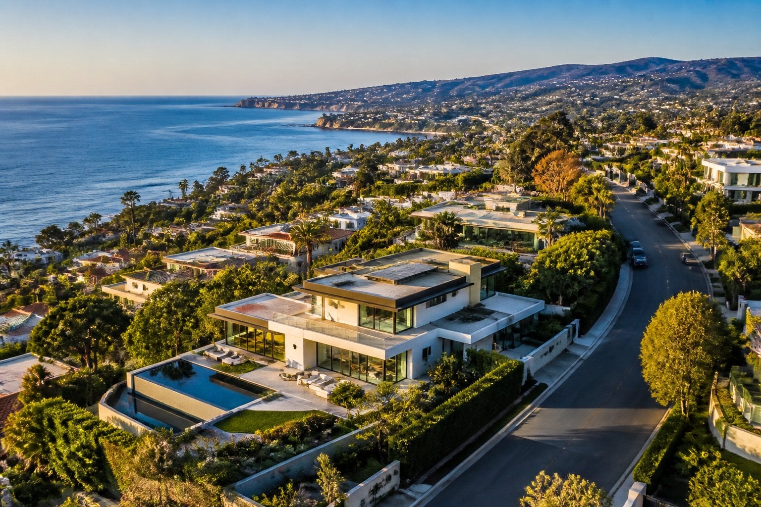 Aerial view of a luxury residential neighborhood with modern homes, trees, hills, and the ocean in the distance.