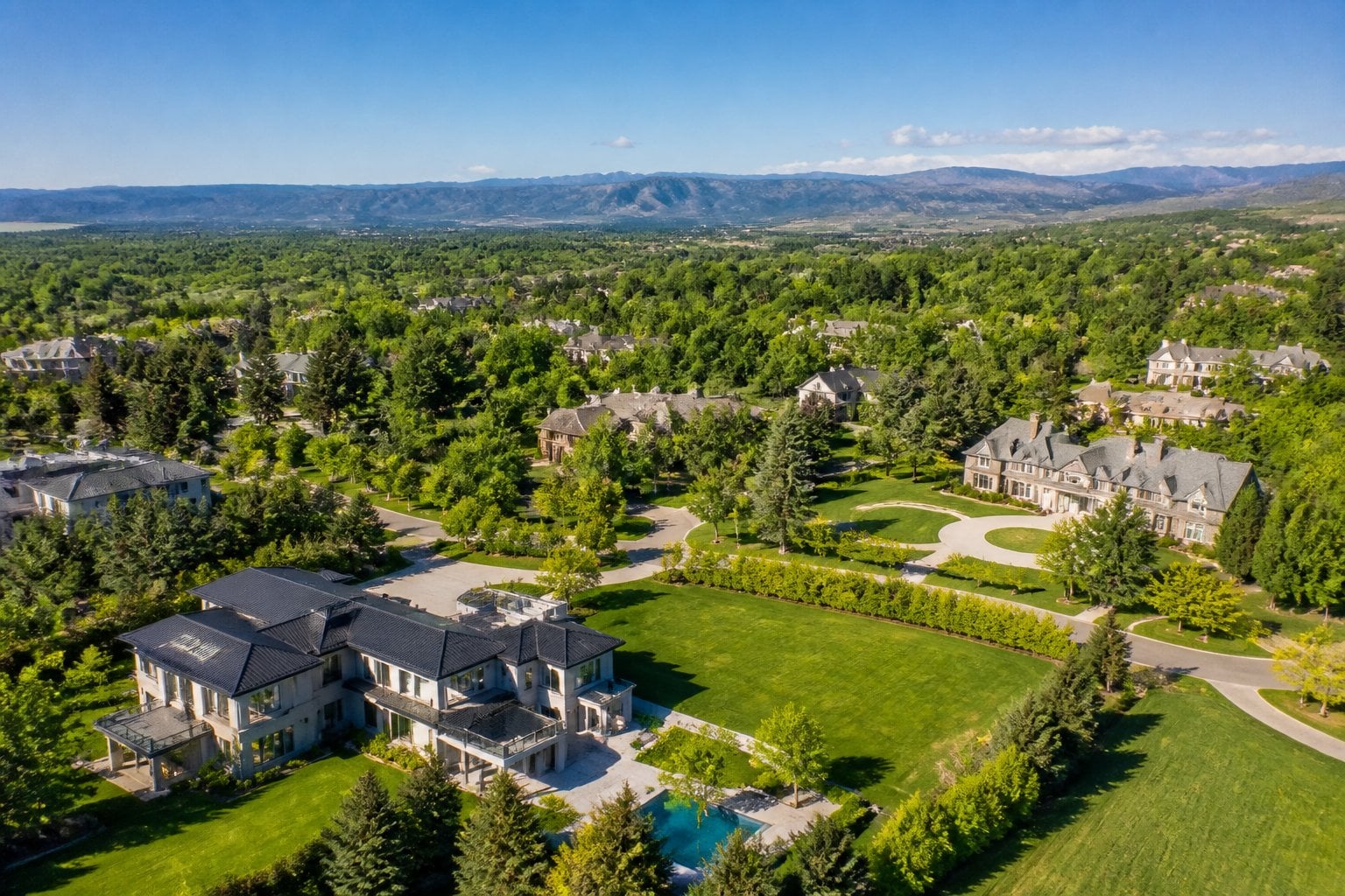 Aerial view of a wealthy residential neighborhood with large houses, green lawns, trees, and hills in the background.
