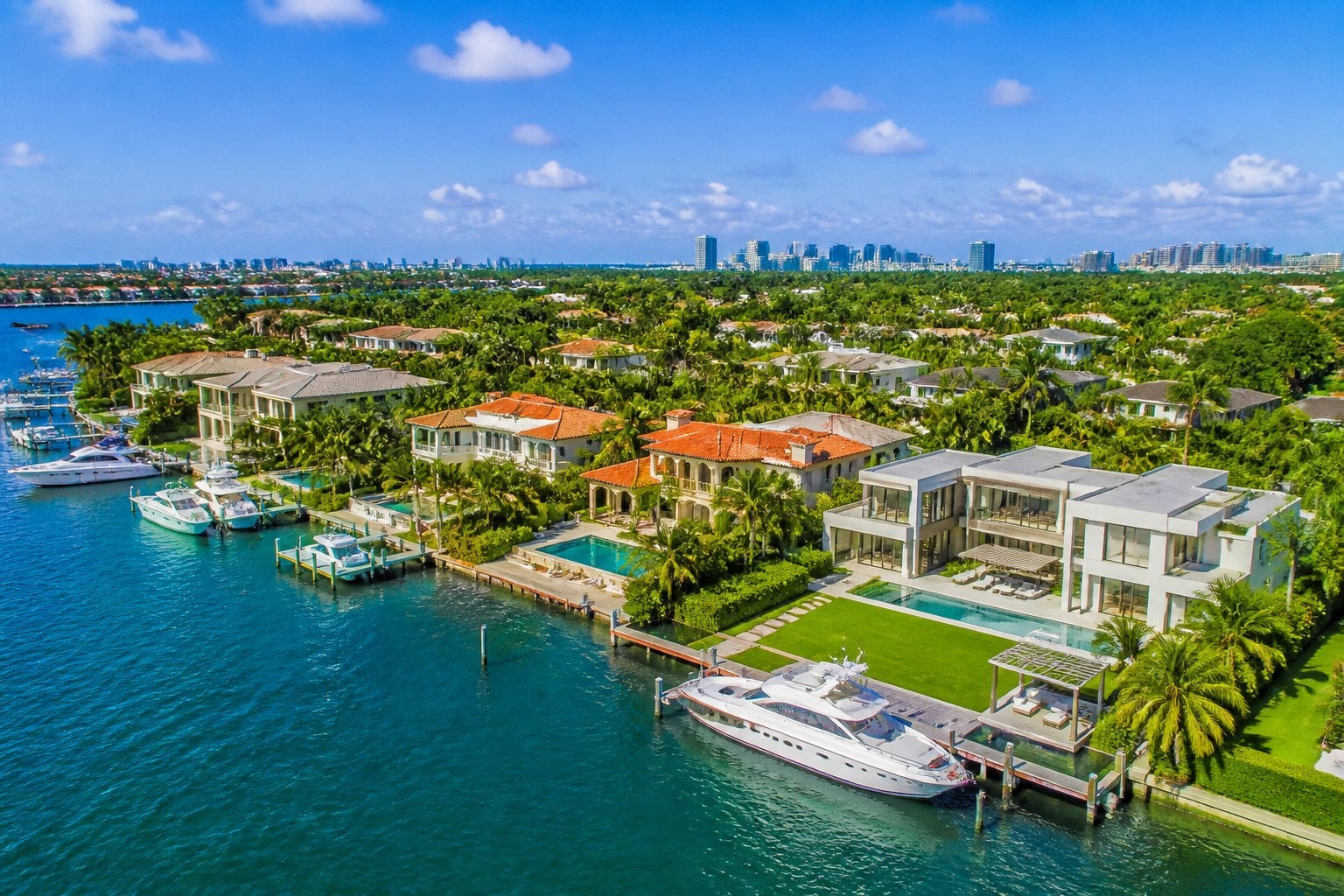 Aerial view of large waterfront mansions with private docks and yachts in a lush, sunny neighborhood.