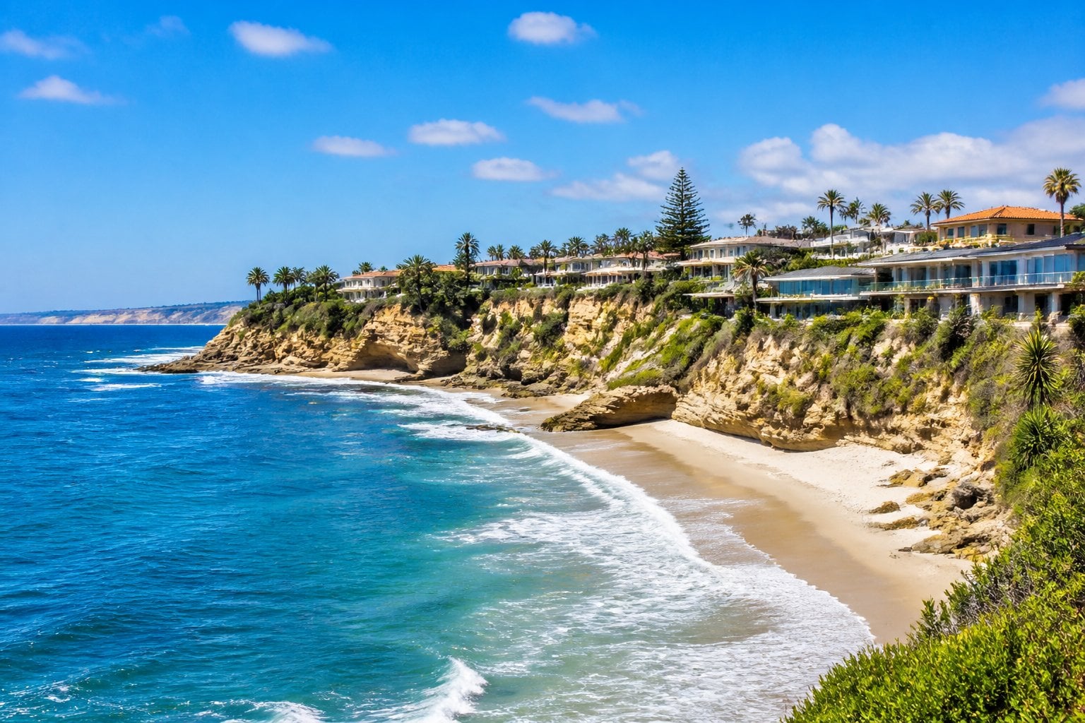 A sunny coastal scene of La Jolla, California with sandy beaches, rocky cliffs, green vegetation, and oceanfront homes overlooking the water.