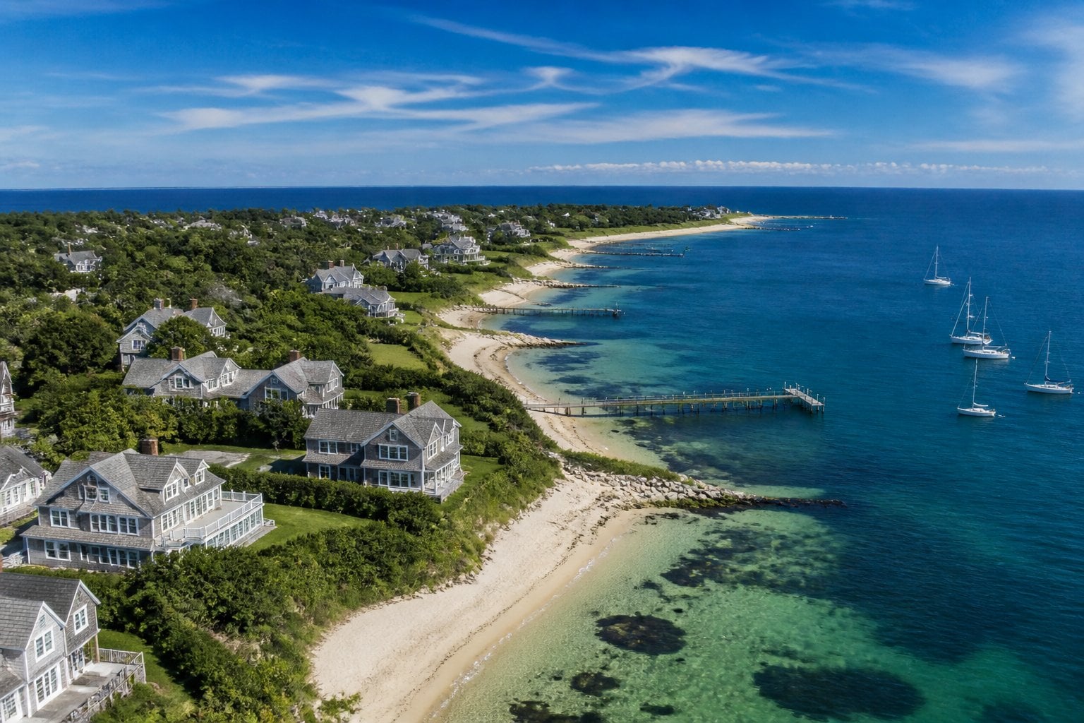 Aerial view of Nantucket coastline with sandy beaches, historic cottages, docks, and sailboats under a sunny sky.
