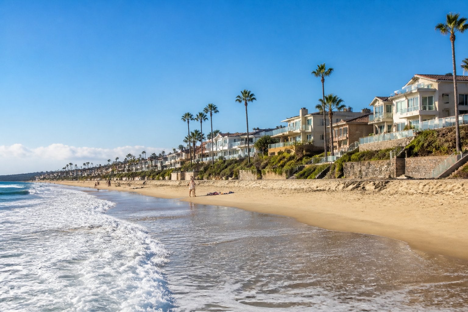 View of a sandy beach with ocean waves, palm trees, and upscale beachfront homes in Manhattan Beach, California.
