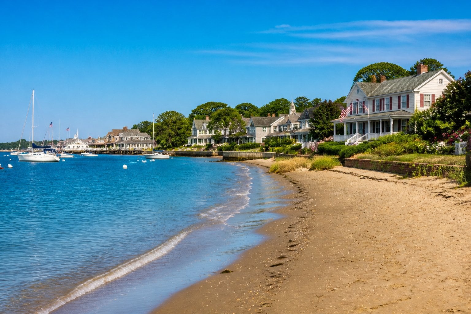 A peaceful beach in Edgartown, Martha's Vineyard with sandy shore, calm ocean waves, classic coastal houses, and boats near the harbor.