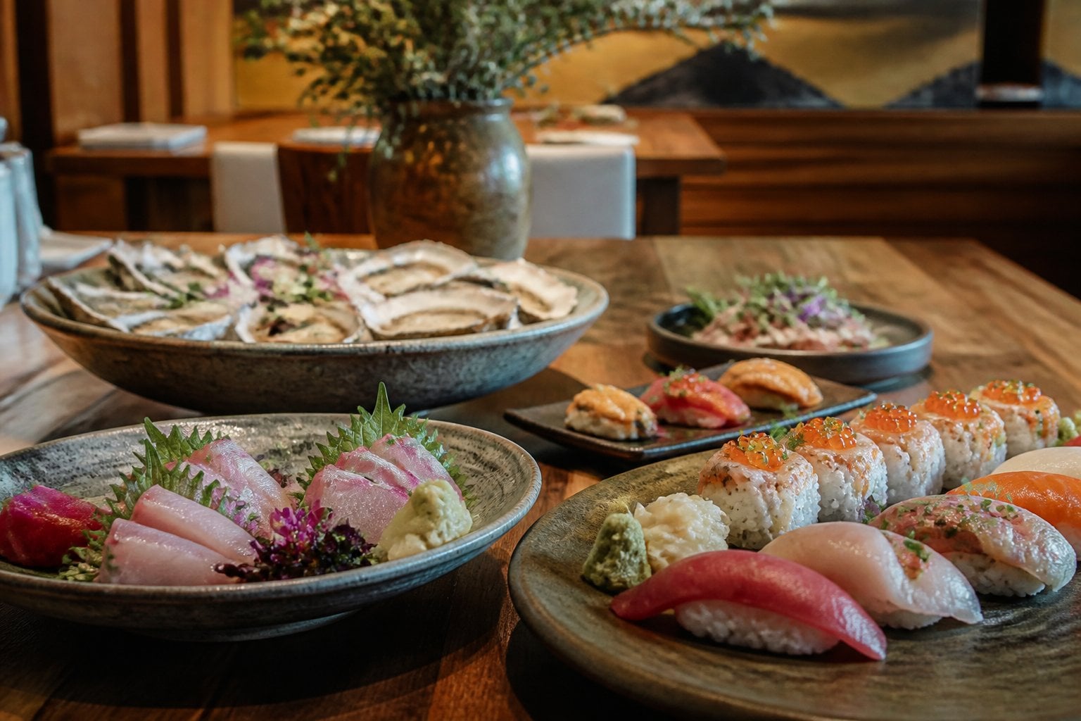A table set with an assortment of fresh seafood dishes including oysters, sushi, and sashimi in a modern restaurant setting.